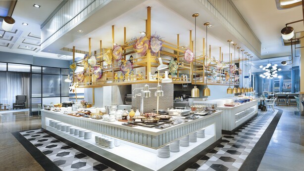 Un amplio vestíbulo de hotel con zona de buffet central, una escalera y una lámpara de araña moderna.