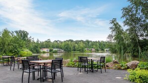 An outdoor patio with tables and chairs, overlooking a lake with houses in the background.