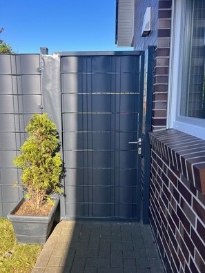 A gated entrance with a small tree in a planter, a brick wall, and a window on the right.