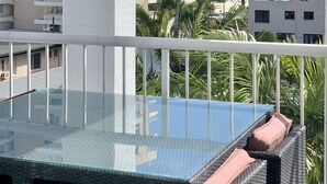 Un balcón con una mesa de vidrio y mobiliario de mimbre, con vista a una playa y edificios altos.