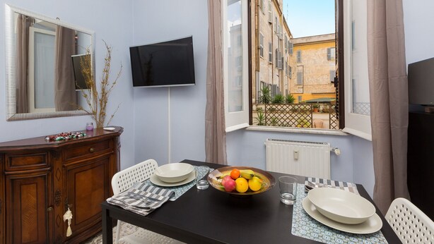A dining area with a table set for two, a fruit bowl, and a television mounted on the wall. - Accogliente (Roma)