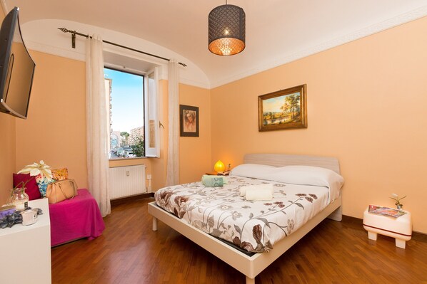 A bedroom with a bed, a window, a bedside table, a lamp, and a painting on the wall. - Accogliente (Roma)