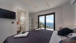 Ein modernes Schlafzimmer mit einem großen Bett, einem an der Wand befestigten Fernseher und einer Glastür, die auf einen Balkon mit Meerblick führt.