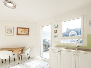 Une cuisine lumineuse avec des meubles blancs, une table à manger en bois et des chaises. Des tableaux encadrés ornent le mur, et une fenêtre offre une vue sur les bâtiments.