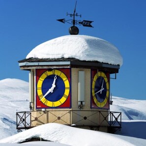 A snow-covered clock tower with two large clocks on its sides, set against a clear blue sky. - At the foot of the Slopes (La Plagne-Tarentaise)