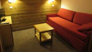 A small, cozy room with a wooden wall, a red sofa, a small table, and two wall-mounted lamps. - At the foot of the Slopes (La Plagne-Tarentaise)