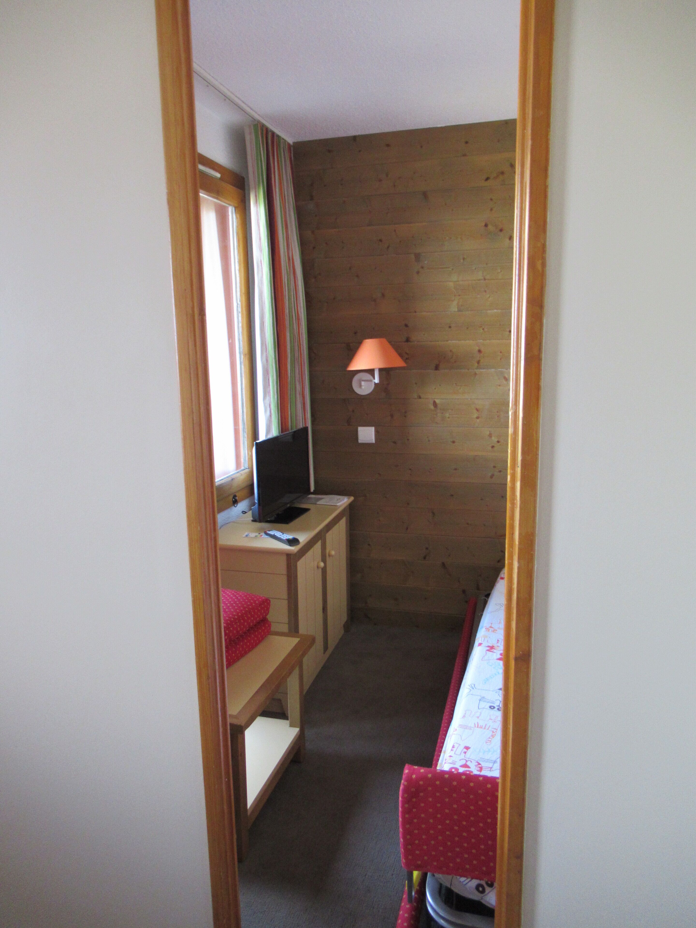 Une pièce étroite aux murs en bois, un bureau avec un ordinateur et une lampe, un lit recouvert d’une couverture rouge et une fenêtre avec des rideaux.