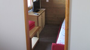 A narrow room with wooden walls, a desk with a computer and a lamp, a bed with a red cover, and a window with curtains. - At the foot of the Slopes (La Plagne-Tarentaise)