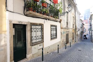 A narrow cobblestone street with a building featuring a green door, barred windows, and a balcony with potted plants.