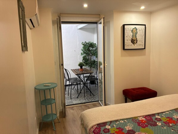 A bedroom with a bed, a small table, a chair, and a red bench. There is a door leading to a balcony with a table and chairs.