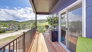 Une terrasse en bois offrant une vue sur une route et des collines, une table avec des chaises et une grande fenêtre.