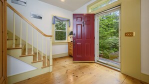 Una escalera de madera lleva a una habitación con una puerta roja, una ventana y vistas al verdor exterior.