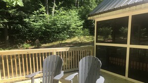 Zwei Adirondack-Stühle auf einer Holzterrasse mit Blick auf einen bewaldeten Bereich und eine überdachte Struktur.