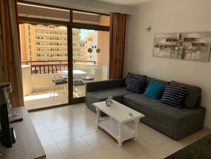 Una sala de estar con un sofá gris, una mesa de centro blanca y un balcón con mesa y sillas.