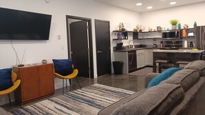 A modern living room with a sofa, a coffee table, and a TV. There is a kitchen area with a microwave, stove, and cabinets. The room has a door and a small trash bin.