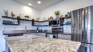 A modern kitchen with a large granite island, stainless steel appliances, and decorative items on shelves.