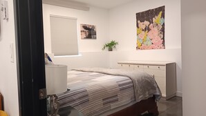 A bedroom with a bed, bedside table, lamp, window with blinds, and a wall with two paintings.