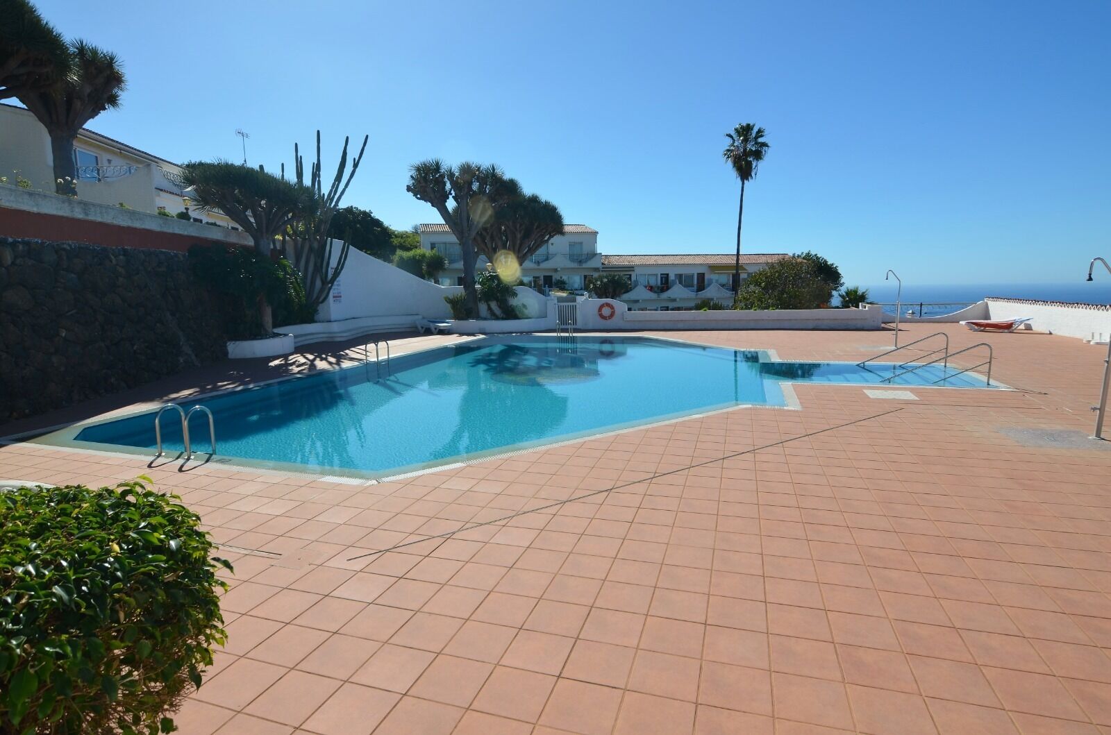 A swimming pool with tiled flooring, surrounded by palm trees and a building with a tiled roof.