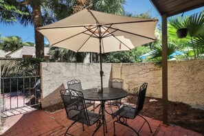 Een terras buiten met een ronde tafel en vier stoelen, een groot beige parasol en een stenen muur op de achtergrond.