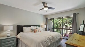 Une chambre à coucher comprenant un lit, des tables de chevet, un ventilateur de plafond, une télévision et un balcon avec vue sur la verdure.