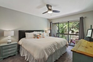 Une chambre à coucher comprenant un lit, des tables de chevet, un ventilateur de plafond, une télévision et un balcon avec vue sur la verdure.