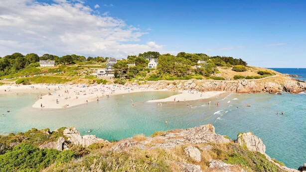 A coastal beach with clear waters, rocky shorelines, and houses on a hillside.