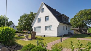 A white two-story house with a sloped roof, a paved driveway, and a well-maintained garden with various plants and a patio area. - Ferienwohnung in Traumhafter Lage am Naturschutzgebiet Salzwiesen (Sylt)