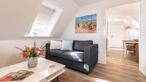 A modern living room with a dark grey sofa, a white coffee table with a vase of flowers, and a painting of a beach scene on the wall. - Ferienwohnung in Traumhafter Lage am Naturschutzgebiet Salzwiesen (Sylt)