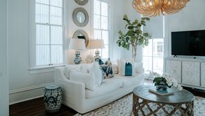 A living room with a white sofa, decorative mirrors, a chandelier, a television, and a large plant.