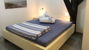 A single bed with a blue mattress and striped blanket, two bedside lamps, and a beach-themed wall art. - Gut Strackholt - Apartment "Seemannsgarn" (Großefehn)