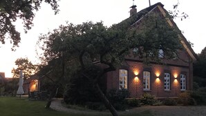 A brick house with a large tree in front, a lawn, and a patio with chairs. - Gut Strackholt - Apartment "Seemannsgarn" (Großefehn)
