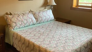 A bedroom with a large bed, a nightstand, a lamp, a framed picture on the wall, and a window with a view of trees. - Hide-away at Yellowstone Archview (Gardiner)