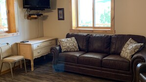 A living room with a brown leather sofa, a wooden desk, a chair, a ceiling fan, and a TV mounted on the wall. - Hide-away at Yellowstone Archview (Gardiner)
