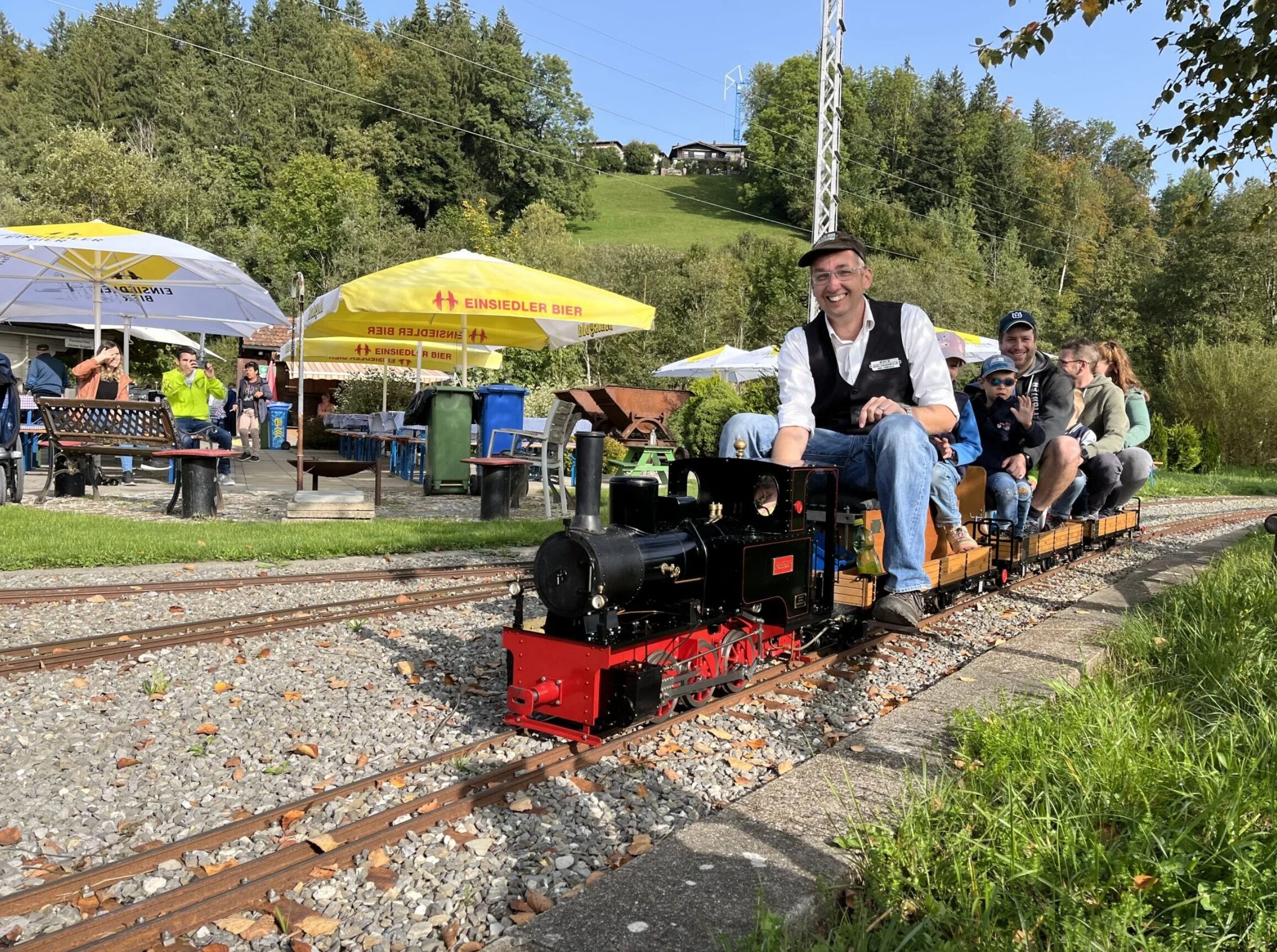Un piccolo treno con passeggeri, binari e una zona erbosa con alberi e un edificio sullo sfondo.