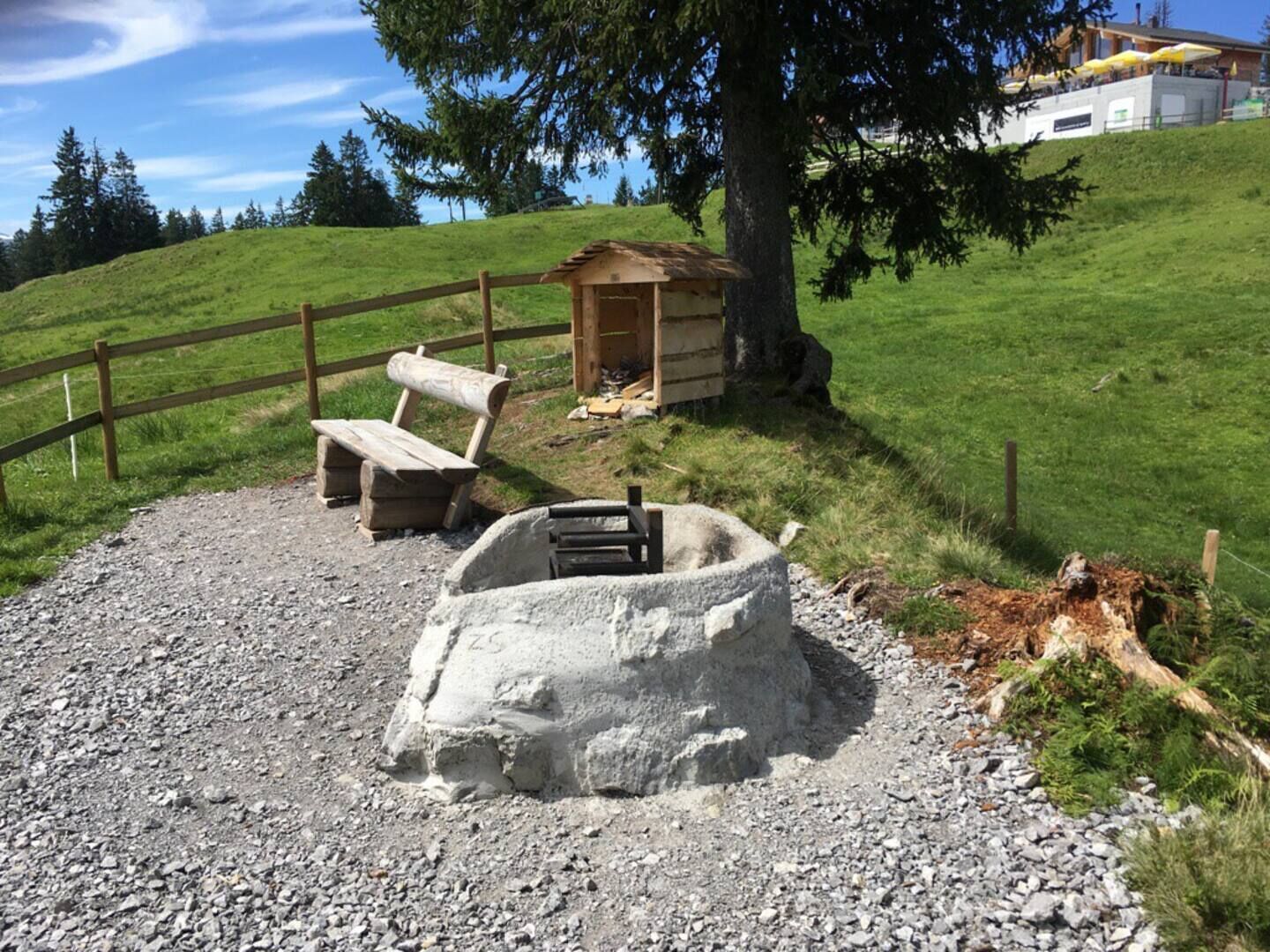 Un espacio al aire libre rústico con un banco de madera y un brasero de piedra, rodeado por una valla de madera y una colina cubierta de césped.