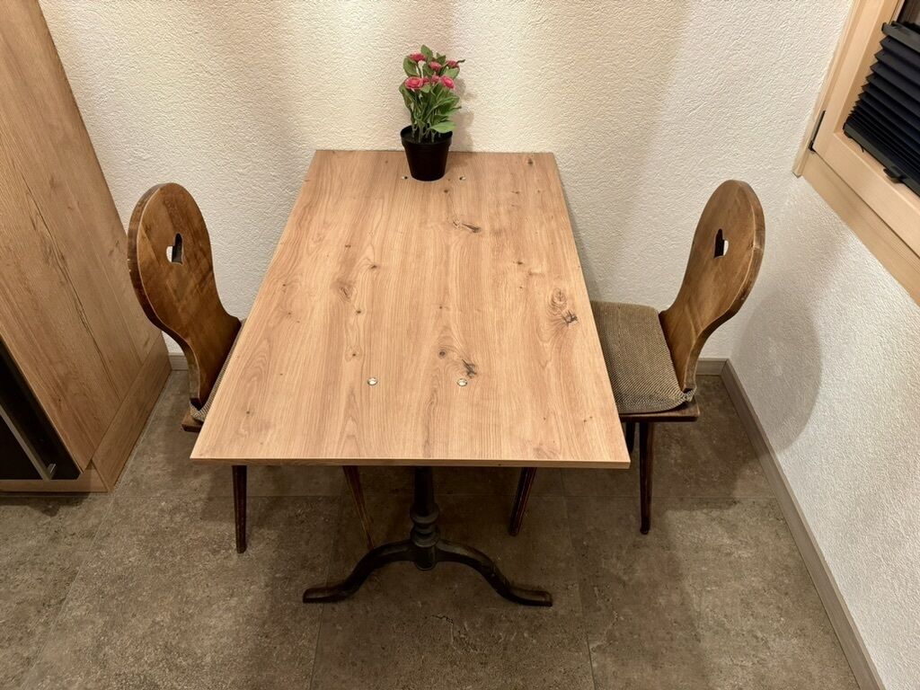 Een houten eettafel met twee stoelen, een potplant en een raam met zonwering.