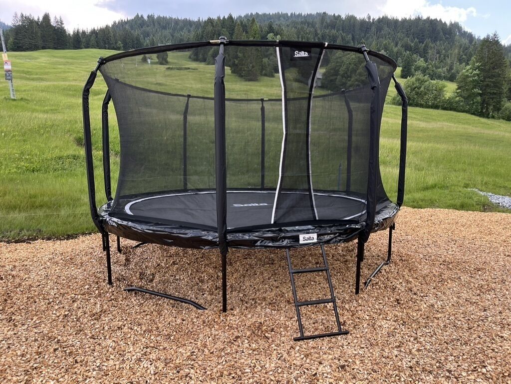 Un trampolín con red de seguridad, sobre una superficie de grava con un campo de césped y árboles al fondo.