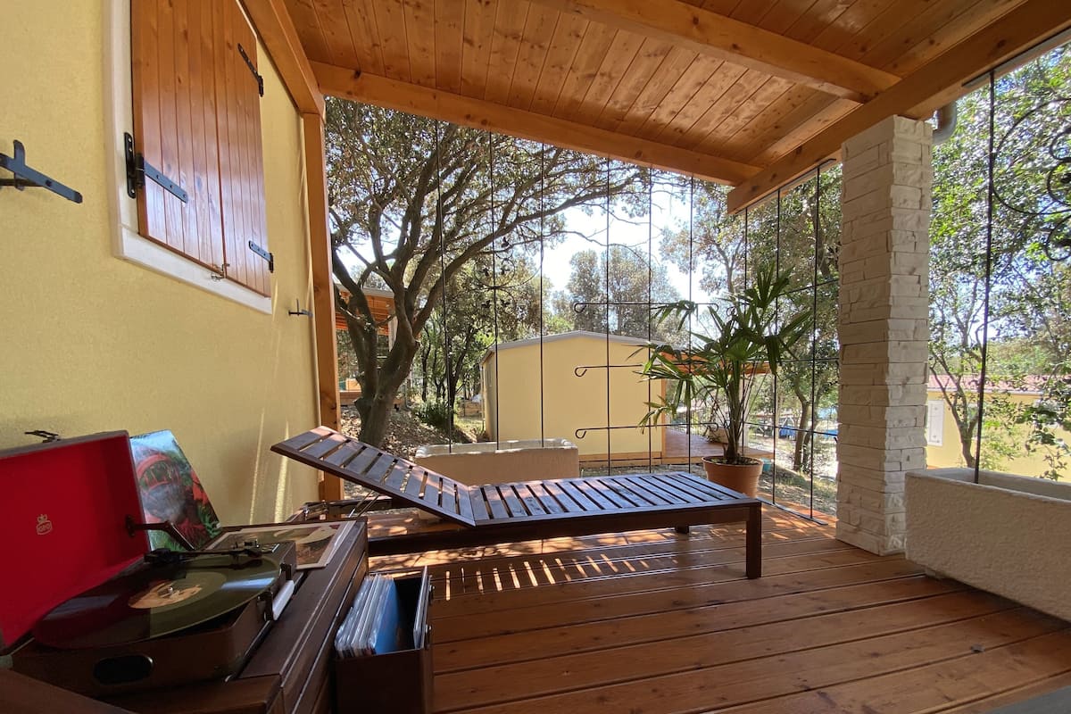 Una terraza de madera con una silla de salón y una mesa, rodeada de macetas con plantas y vistas a árboles.