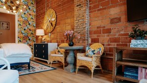 A cozy room with a brick wall, a round table, a wicker chair, a white sofa, a dark-colored dresser, and a bookshelf. - THE PINK ROOM @emilysontheisland (Galveston)