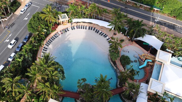 An aerial view of a resort with a central swimming pool surrounded by palm trees, a covered seating area, and a road with cars. - 2 and 3 Bedroom Apartments within Crown Towers (Surfers Paradise)
