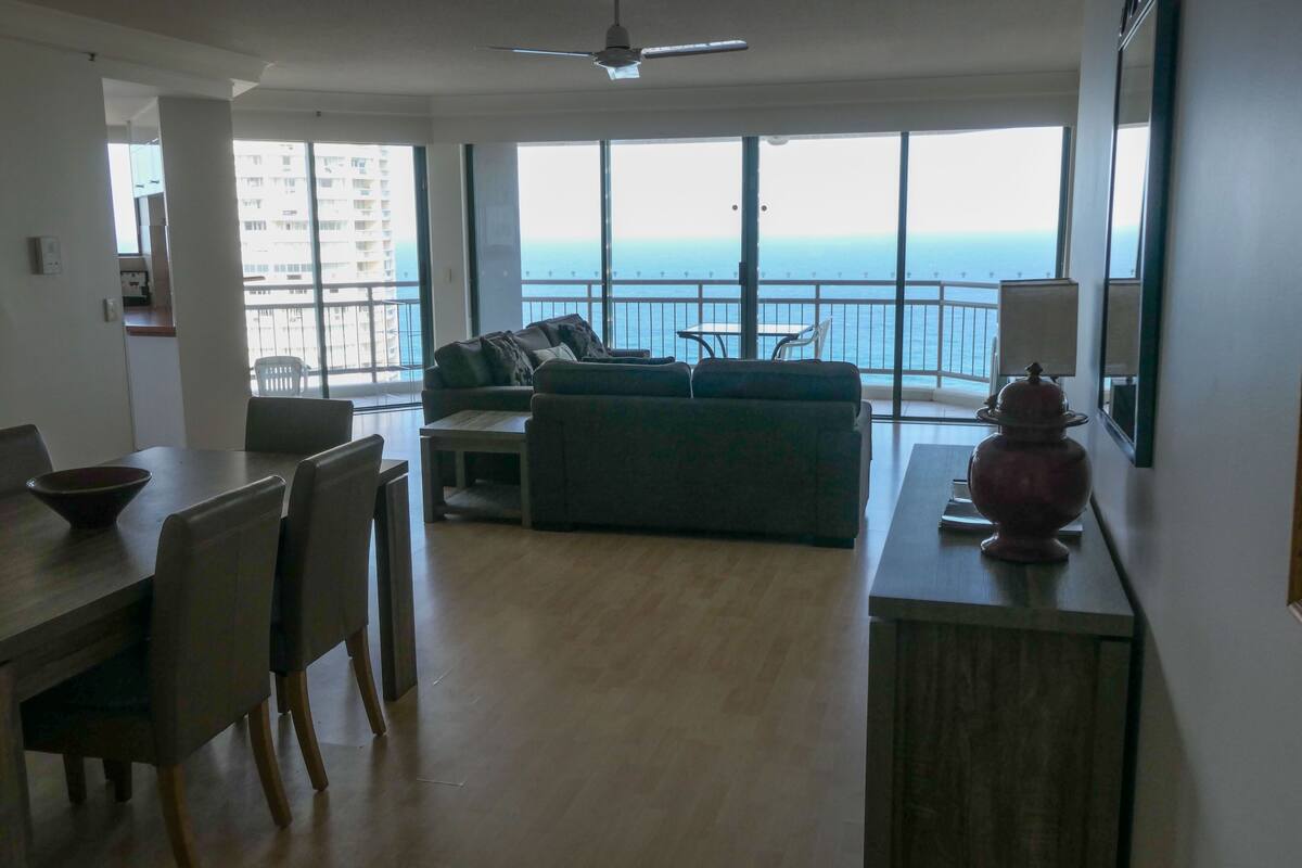 Amplia zona de estar con balcón vistas al mar, una mesa de comedor con sillas y un sofá con cojines.