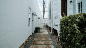 A narrow alleyway with a stone pathway flanked by white buildings, a hedge on one side, and a bicycle parked in the distance.