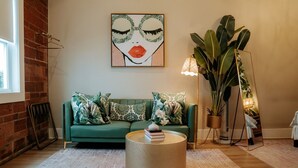 A green sofa with patterned cushions, a round coffee table, a large plant, and a painting on the wall.