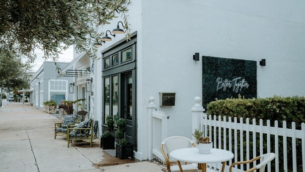 A white building with a black sign reading 'Better Together,' outdoor seating with wicker chairs and a table, and a white picket fence.
