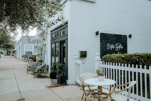 Un bâtiment blanc avec une enseigne noire sur laquelle on peut lire « Mieux ensemble », des tables et des chaises en osier disposées à l’extérieur, et une clôture blanche faite de piquets.