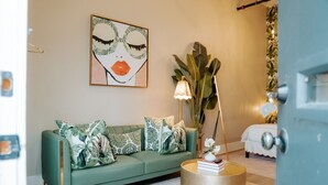 A green sofa with patterned cushions, a round gold side table, a large plant, and a painting on the wall.