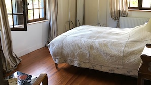 Ein Schlafzimmer mit Holzboden, einem Bett mit Tagesdecke, einem Sessel, einem Heizkörper und einem Fenster mit Vorhängen.