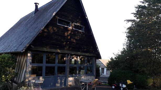 Ein Holzhaus mit einem steilen Satteldach, großen Fenstern und einer Terrasse mit Gartenmöbeln.