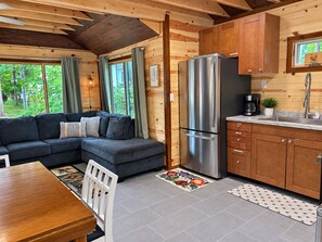 A modern log cabin interior with a sectional sofa, a stainless steel refrigerator, wooden cabinets, and a kitchen counter.