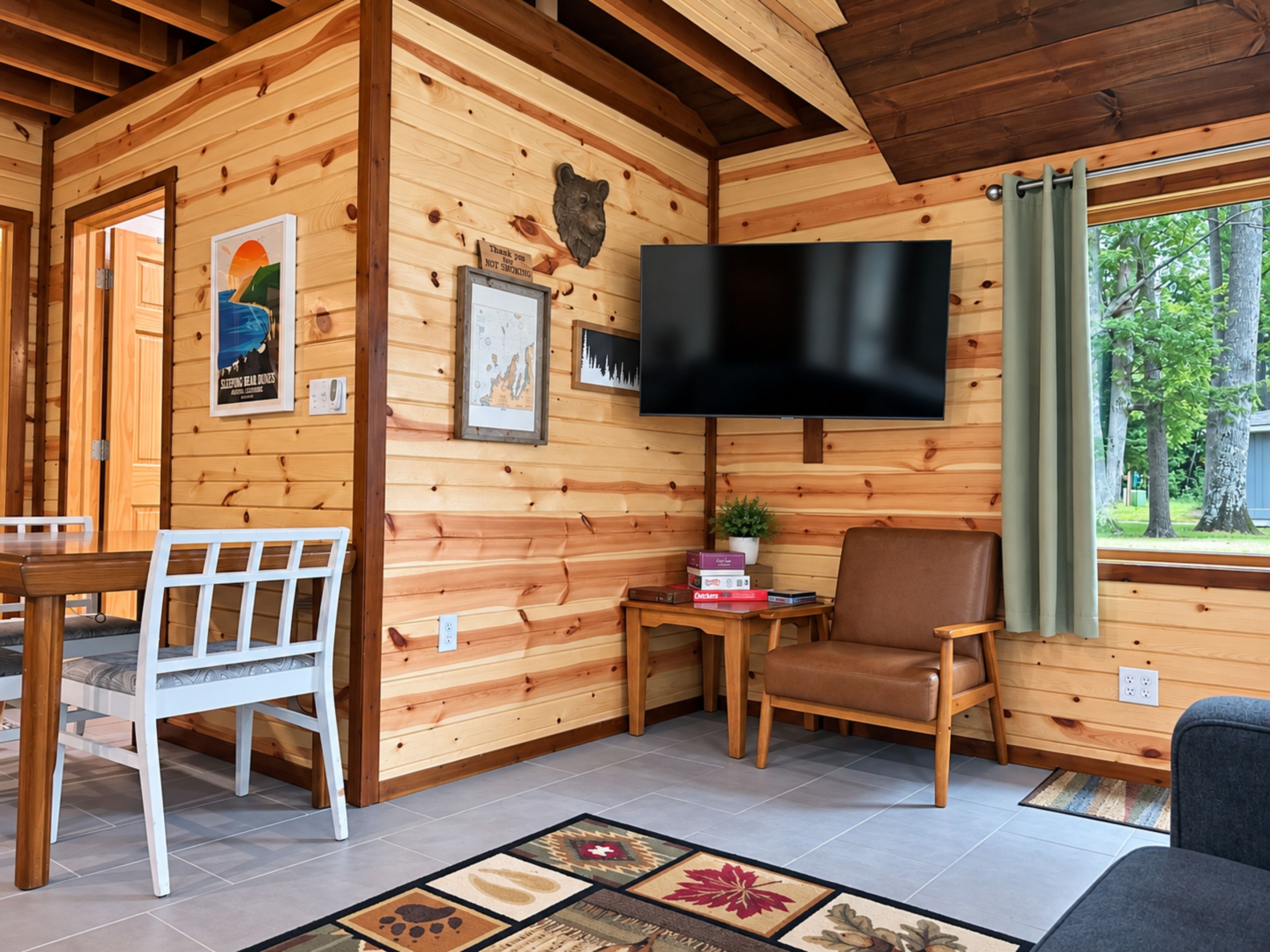 A wooden cabin interior with a television, a dining table, chairs, and a window with a view of trees.